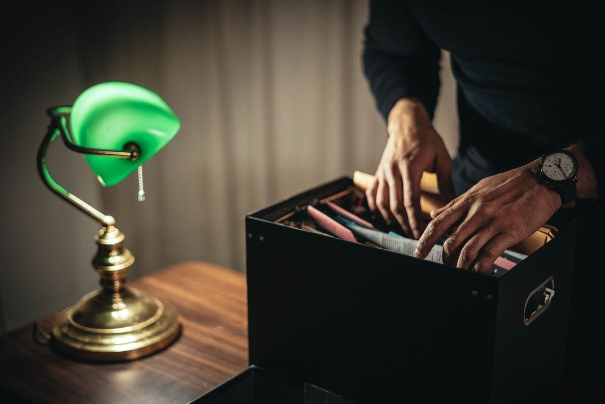 Une main dans une mallette pleine de papiers, une lampe verte sur une table Une main dans une mallette pleine de papiers, une lampe verte sur une table