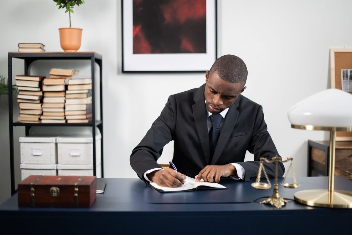 Homme en costume noir écrivant à un bureau avec une balance, un livre et une lampe. Tableau rouge au mur. Étagère avec livres Homme en costume noir écrivant à un bureau avec une balance, un livre et une lampe. Tableau rouge au mur. Étagère avec livres