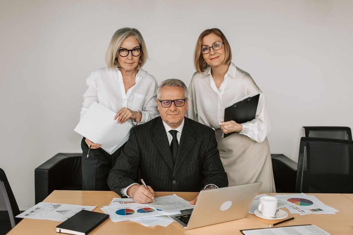 Homme en costume écrit à un bureau avec deux femmes en blouses devant des graphiques et un ordinateur
