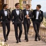 Quatre hommes élégants en smoking noir et nœud papillon marchant sur une terrasse lors d'un mariage prestigieux.