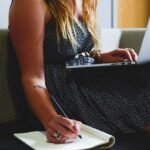 Une femme écrivant dans un carnet tout en utilisant un ordinateur portable